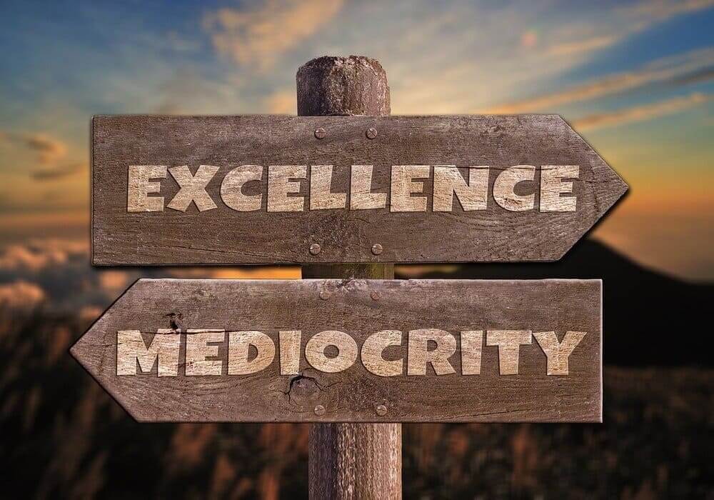 A wooden signpost against a sunset sky, pointing in two directions labeled 'Excellence' and 'Mediocrity,' symbolizing the choices we face in life. This image reflects the idea that security often lures us into mediocrity, while striving for excellence demands stepping beyond our comfort zones